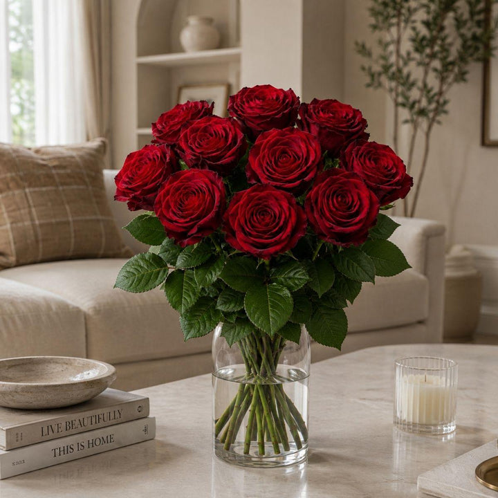 A bouquet of 12 deep red roses in a clear glass vase, placed on a marble coffee table in a cozy, modern living room setting.