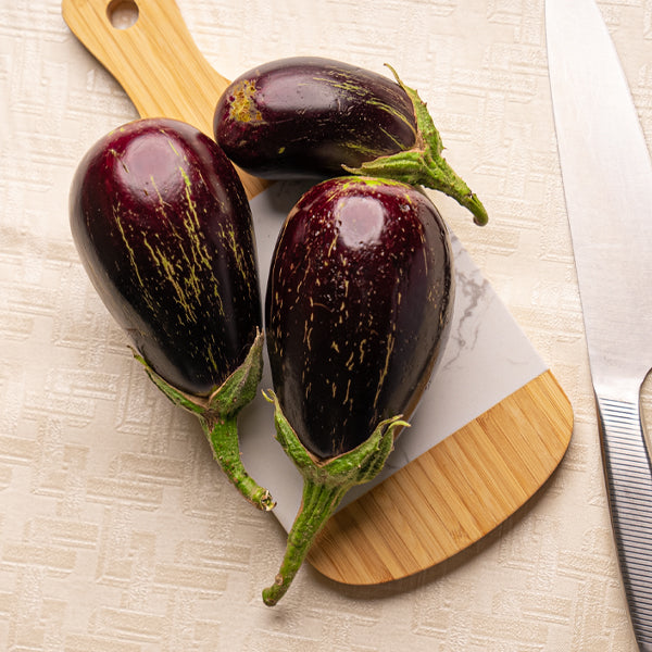 Fresh organic eggplants from Naqwat Al Nana, 1 kg pack, showing glossy purple vegetables with green stems on a white background.