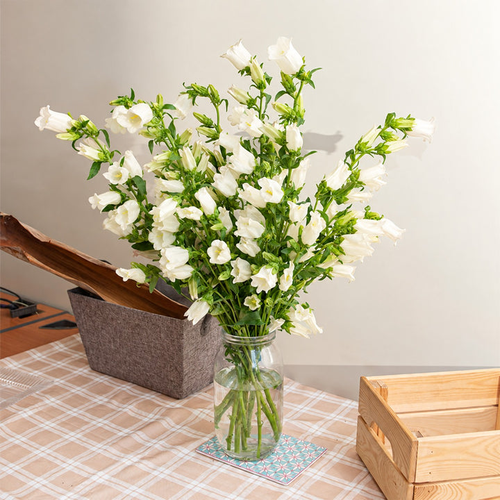 A bouquet of white bell-shaped flowers arranged in a clear glass jar vase, placed on a checkered tablecloth beside a gray felt box and a wooden crate.