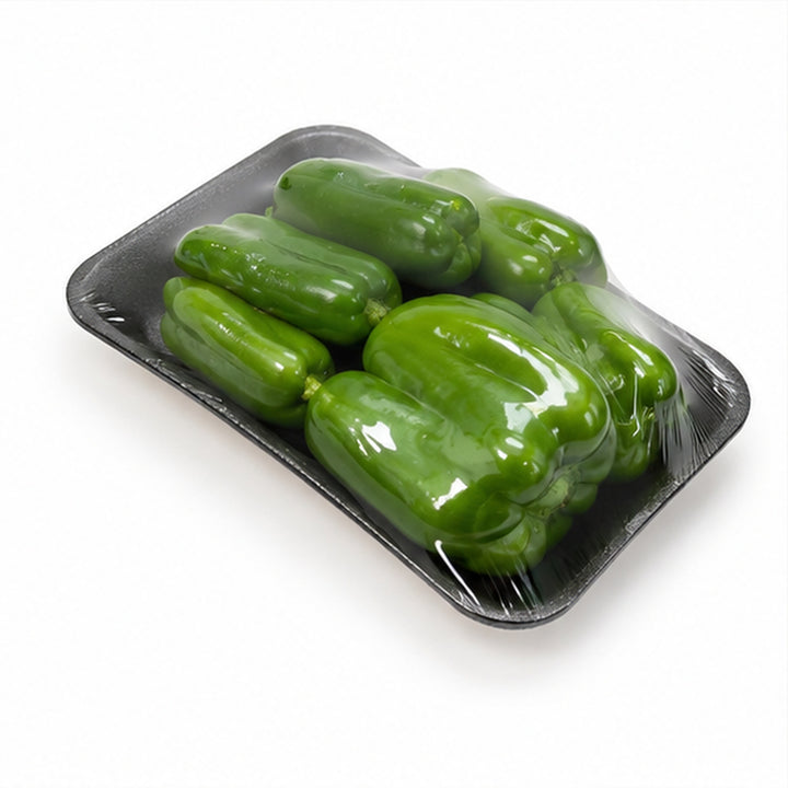 A black plastic tray holding several glossy green bell peppers, covered with clear plastic wrap, on a white background.